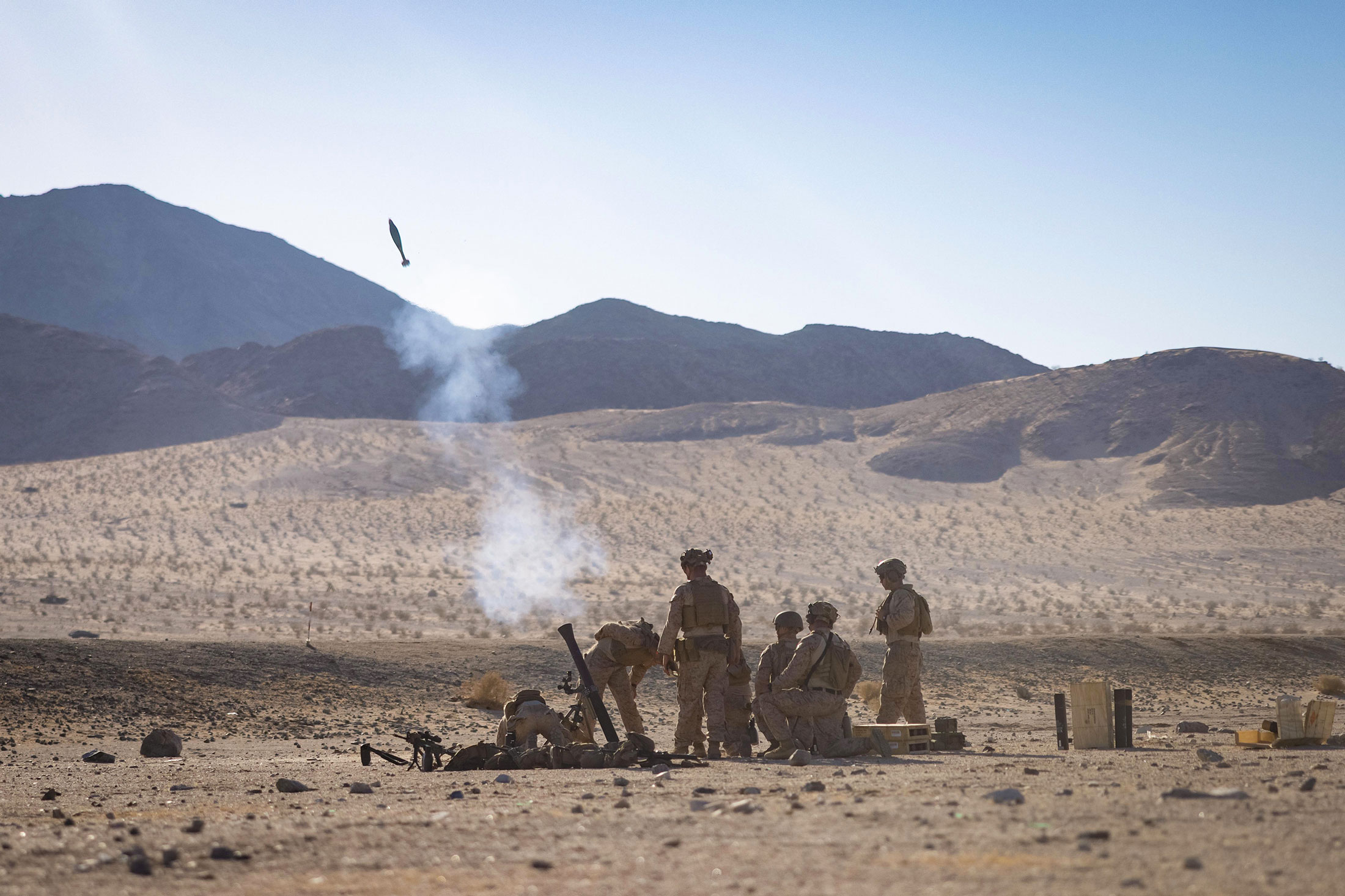 A team of U.S. Marines is seen operating and firing an M252 81 mm mortar system in a desolate, rocky desert environment, with the mortar round visible high in the air just after launch and a puff of smoke rising from the tube.