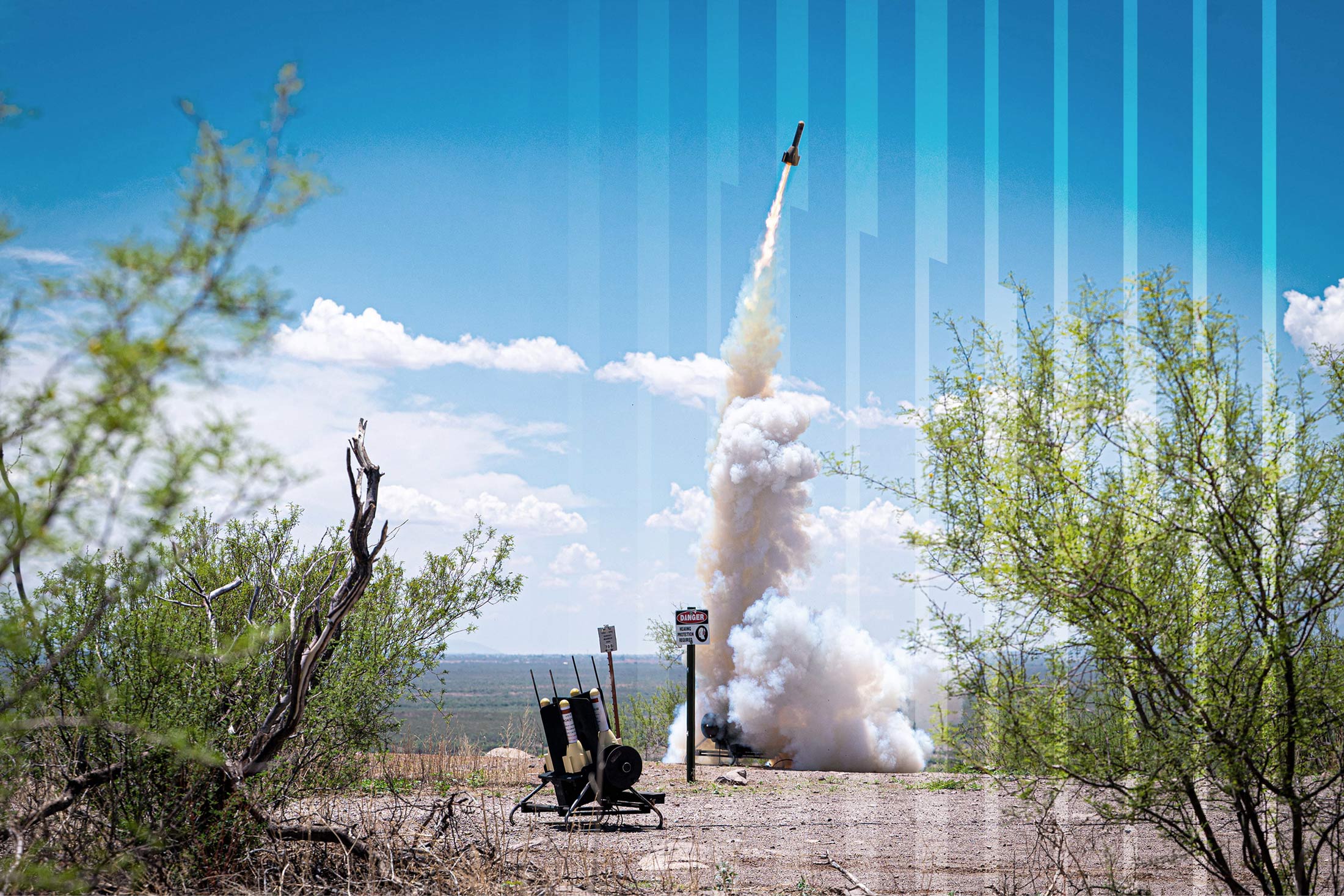 A surface-to-air rocket simulator, known as "Smokey Sam," is shown launching from its ground mount, leaving a thick trail of white smoke that rises high into the clear, blue sky with scattered clouds. The launch system is visible in the foreground, surrounded by desert scrub and a warning sign.