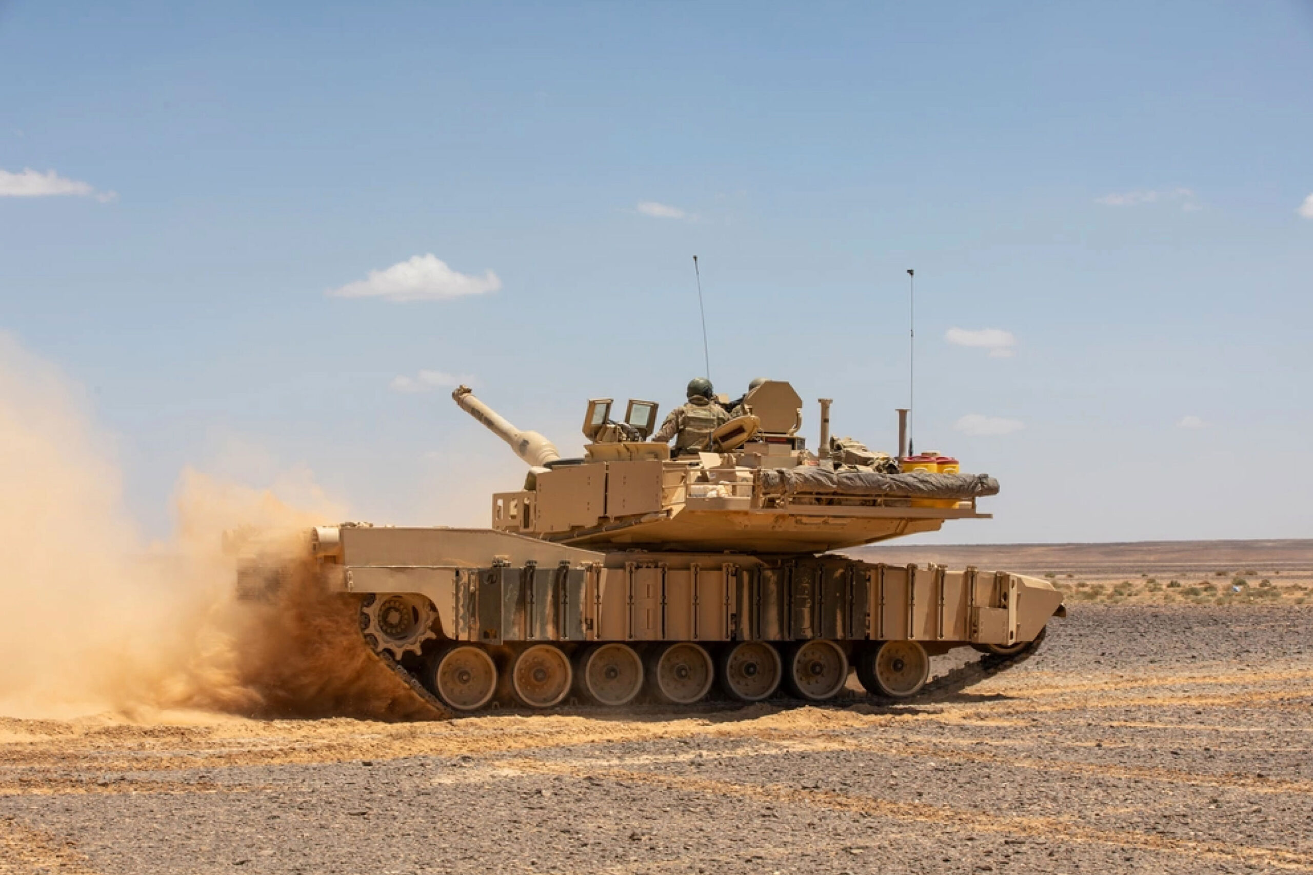 Desert tan U.S. Army M1A1 Abrams tank driving through a dry, rocky field, kicking up a cloud of dust behind it.